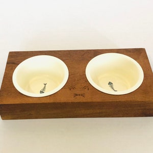 May include: A wooden cat bowl with two cream-colored bowls. Each bowl has a gray fishbone design. The wooden base is rectangular and has a cat face design. The wood is a dark brown color.