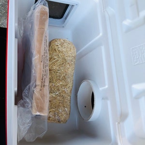 以下が含まれることがあります： 赤い縁取りのある白いクーラーボックスが開いており、プラスチックで包まれた長方形の物体、円筒形の藁のような物体、白いPVCパイプが入っています。 クーラーボックスには通気孔があり、「Warning」の文字が見えます。