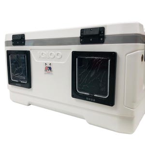 May include: White Igloo cooler with two black pet door inserts. The cooler has a gray trim and black latches. An American flag sticker is on the front. The cooler has a black handle on the side.
