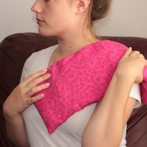 May include: A pink fabric heating pad is being held against a person's shoulder. The heating pad is made of a soft, fuzzy material and has a subtle pattern.