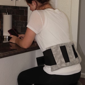 May include: A gray and black heating pad is worn around the lower back. The pad has a rectangular shape with three gray sections and two black sections. The person is wearing a white t-shirt and black leggings.
