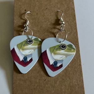 May include: A pair of silver dangle earrings with a light blue guitar pick shaped charm featuring a green frog wearing a red and blue bow tie.