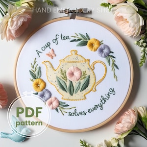 Puede incluir: Un aro de bordado blanco con un diseño cosido a mano de una tetera, flores y hojas. El texto "A cup of tea solves everything" está cosido con hilo verde alrededor de la tetera. El aro está sobre un fondo blanco.