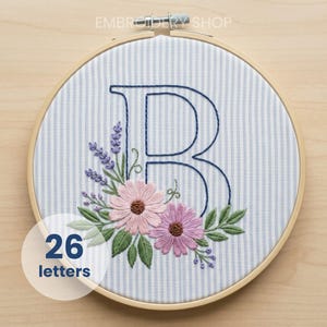 以下が含まれることがあります： 紺色の糸で刺繍された文字「B」、ピンクと紫の花と緑の葉で囲まれています。 デザインはストライプの生地にあり、木製の刺繍枠にセットされています。 テキスト「26 letters」は白い円の中にあります。