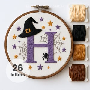 May include: A Halloween-themed embroidery hoop featuring a purple letter "H" with a black witch hat, spider, and spiderwebs. The design includes orange and purple stars. The text "26 letters" is in a white circle. Spools of thread are to the right.