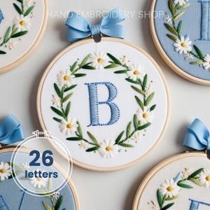 Puede incluir: Arte de aro bordado con fondo blanco, letra "B" azul y una corona floral de margaritas blancas y hojas verdes. Una cinta azul está atada en la parte superior. El texto "HAND EMBROIDERY SHOP" está en la parte superior.