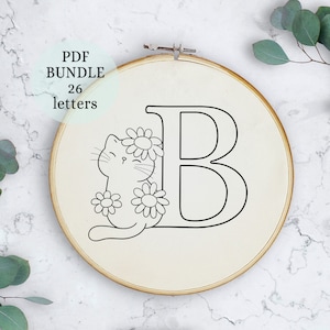 May include: A black and white embroidery design of the letter B with a cat and flowers. The design is on a white fabric in a wooden embroidery hoop.