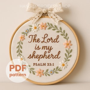 May include: A circular embroidery hoop with the text "The Lord is my shepherd" and "Psalm 23:1". The design includes a floral wreath with peach, blue, and white flowers. A lace bow adorns the top. The hoop is a PDF pattern.