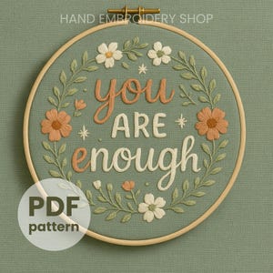 Puede incluir: Arte de aro bordado con las palabras "you are enough" en cursiva. El diseño presenta flores naranjas y blancas, hojas verdes y pequeños acentos de estrellas sobre un fondo verde salvia. El aro es de madera.