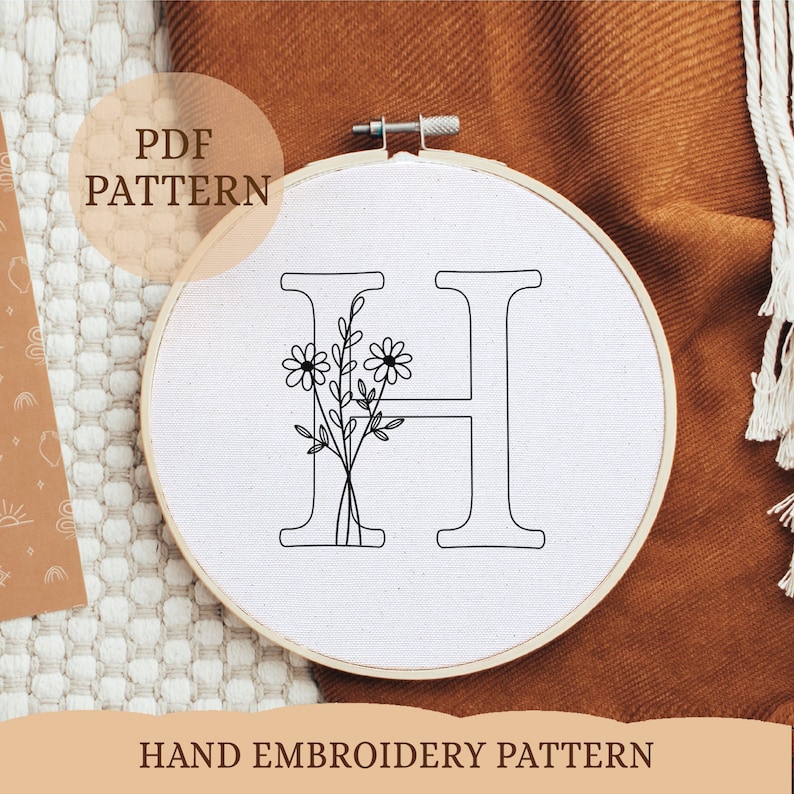 May include: A black and white hand embroidery pattern of the letter H with a floral design. The pattern is on a white fabric background and is displayed in a wooden embroidery hoop.