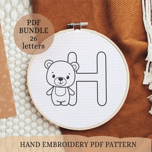 May include: A black and white hand embroidery pattern of a teddy bear holding the letter H. The pattern is on a white fabric in a wooden embroidery hoop. The text "PDF BUNDLE 26 letters" is in a white box with a black outline.
