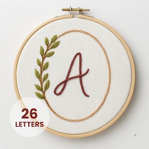 Puede incluir: Arte de aro bordado con la letra "A" en hilo burdeos, rodeada por una corona cosida de hojas verdes y un borde beige. El texto "26 LETTERS" está en un círculo blanco. El aro de madera es visible.