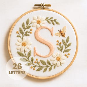 Puede incluir: Arte de aro bordado con la letra "S" en melocotón, rodeada de margaritas, hojas verdes y una abeja. El texto "26 LETTERS" está en un círculo blanco. El bordado está en un aro de madera.