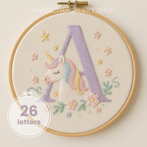 Op de afbeelding: Geborduurde hoepelkunst met een eenhoorn en de letter "A" in lavendel. De eenhoorn heeft een regenboogmanen en een gouden hoorn. Het ontwerp bevat bloemen en sterren, alles in een houten hoepel. De tekst "26 letters" is ook inbegrepen.