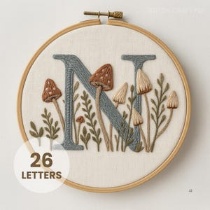 May include: Embroidered hoop art with the letter "N" in grey, surrounded by brown and beige mushrooms and green foliage. The design is set against a cream-coloured fabric, framed by a wooden hoop. The text "26 LETTERS" is in a white circle.