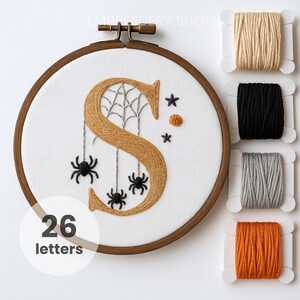 May include: An embroidery hoop with a stitched letter "S" in gold, featuring a spider web and hanging spiders. The design includes small purple stars and an orange pom-pom. The text "26 letters" is in a white circle. Spools of thread in beige, black, gray, and orange are to the right.