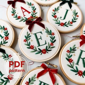 Puede incluir: Aro de bordado con letras A, E, J y K, cada una rodeada por una corona de hojas verdes y flores rojas. Las letras son rojas y verdes. Cada aro tiene un lazo de terciopelo rojo o verde. El texto "PDF pattern" es visible.