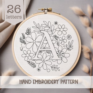 May include: A black and white hand embroidery pattern featuring the letter A surrounded by a wreath of flowers and leaves. The pattern is on a white fabric background and is labeled "HAND EMBROIDERY PATTERN".