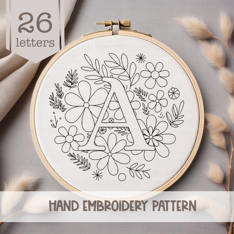 May include: A black and white hand embroidery pattern featuring the letter A surrounded by a wreath of flowers and leaves. The pattern is on a white fabric background and is labeled "HAND EMBROIDERY PATTERN".