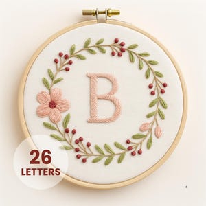 May include: Embroidered hoop art featuring the letter "B" in pink, surrounded by a floral wreath with green leaves and red berries. A pink flower is on the left side. The text "26 LETTERS" is in a white circle at the bottom.