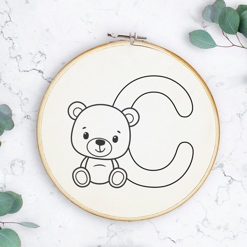 May include: A white embroidery hoop with a black line drawing of a teddy bear sitting next to the letter C.