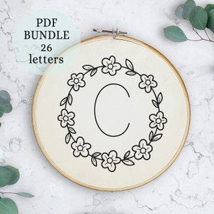 May include: A black and white embroidery design of the letter C surrounded by a wreath of simple flowers and leaves. The design is on a white fabric background in a wooden embroidery hoop.