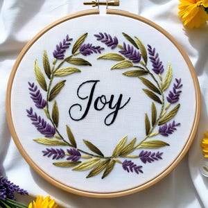 May include: Embroidered hoop art featuring a wreath of purple and green flowers with the word "Joy" in the centre.