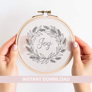 May include: A white embroidery hoop with a black line drawing of a wreath with the word "Joy" inside. The hoop is being held by two hands.