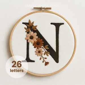 May include: Embroidered letter "N" in dark brown, adorned with floral accents in shades of brown and beige. The design is set within a wooden embroidery hoop. The text "26 letters" is in a white circle. The shop name "Embroidery Shop" is at the top.