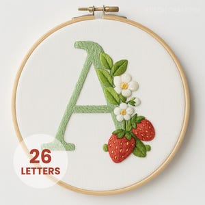 Può includere: Lettera "A" ricamata in verde chiaro, ornata da fiori bianchi, foglie verdi e fragole rosse. Il ricamo è posto in un cerchio di legno. Il testo "26 LETTERS" è in un cerchio bianco.