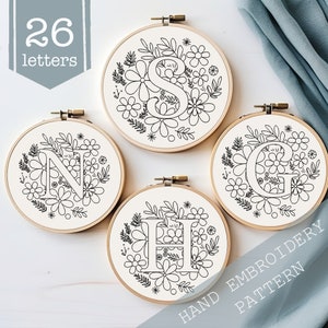 May include: Four embroidery hoop patterns featuring the letters N, S, H, and G surrounded by floral designs. The hoops are on a white surface with the text "HAND EMBROIDERY PATTERN" at the bottom right corner.