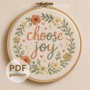 Op de afbeelding: Geborduurd kunstwerk in een houten ring met de woorden "choose joy" in sierlijke letters. Het ontwerp bevat een bloemenkrans in perzik, groen en crème. Een decoratief stuk voor in huis.