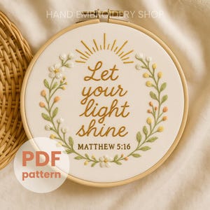 Op de afbeelding: Geborduurde borduurring met de tekst "Let your light shine" in gouddraad. Het ontwerp bevat een bloemenkrans met witte, gele en perzik kleurige bloemen en groene bladeren. De tekst "Matthew 5:16" staat eronder. De woorden "PDF pattern" staan in een cirkel.