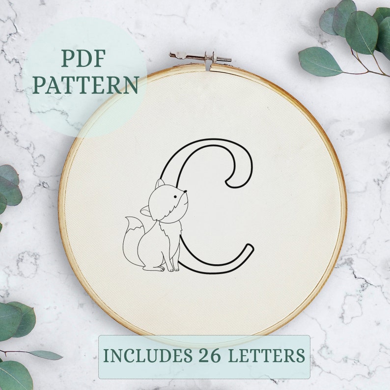 May include: A black and white embroidery pattern of the letter C with a fox sitting next to it. The pattern is on a white fabric in a wooden embroidery hoop. The text "PDF PATTERN" is in a circle above the hoop and the text "INCLUDES 26 LETTERS" is below the hoop.