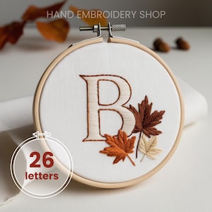 May include: A hand-embroidered letter "B" in a wooden hoop, featuring autumn-colored leaves. The embroidery is on white fabric, with the text "HAND EMBROIDERY SHOP" at the top. A circular graphic with the text "26 letters" is in the lower left.