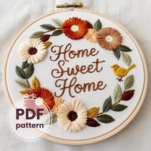 May include: Embroidery hoop with a floral wreath design featuring orange, yellow, and brown flowers. The text "Home Sweet Home" is embroidered in the centre of the hoop.