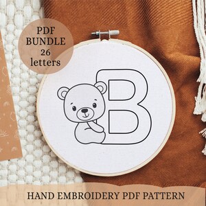 May include: A black and white line drawing of a bear sitting next to the letter B. The image is on a white fabric background in a wooden embroidery hoop. The text "PDF BUNDLE 26 letters" is in a white box with a black outline.