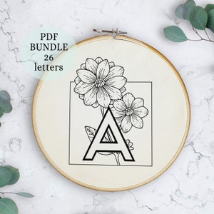 May include: A black and white embroidery pattern of the letter A with two stylized flowers. The pattern is in a square frame on a beige background. The text "PDF BUNDLE 26 letters" is in a white box in the upper left corner.