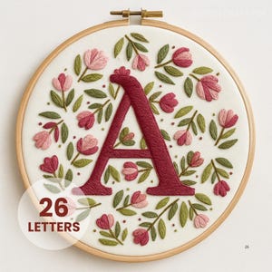 Peut inclure: Broderie d'un lettrage "A" bordeaux, entouré de motifs floraux roses et rouges avec des feuilles vertes, dans un cercle à broder en bois. Le texte "26 LETTERS" est dans un cercle blanc en bas à gauche.