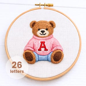 May include: Embroidered teddy bear hoop art. The bear is brown with a pink sweater featuring a red letter "A" and blue pants. The text "26 letters" is in a white circle in the lower left corner. The embroidery is set in a wooden hoop.
