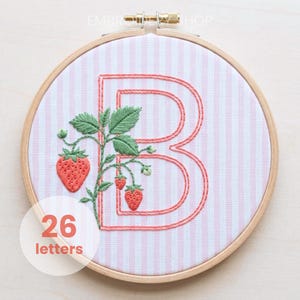 May include: Embroidered letter "B" with a strawberry plant design, framed in a wooden hoop. The background is pink and white striped fabric. The text "26 letters" is in a white circle.