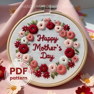 May include: A white embroidery hoop with a red, pink, and green floral design. The hoop has the text "Happy Mother's Day" embroidered in the center. The hoop is on a pink surface. PDF pattern.