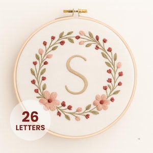 May include: Embroidered hoop art featuring the letter "S" in beige thread, surrounded by a floral wreath of pink and red flowers and green leaves. The text "26 LETTERS" is in a white circle in the lower left corner. The embroidery is set in a wooden hoop.