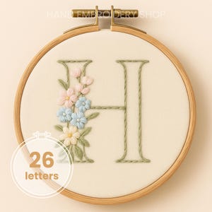 Könnte beinhalten: Gestickter Buchstabe "H" in einem Holzreifen. Der Buchstabe ist mit hellgrünem Faden gestickt und mit rosa, blauen und cremefarbenen Blumenstickereien verziert. Der Text "26 Buchstaben" befindet sich in einem weißen Kreis unten.