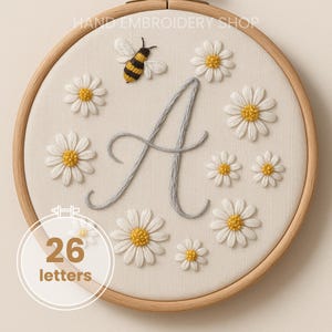 May include: Embroidered hoop art with a daisy and bee design and the letter "A". The daisies have white petals and yellow centres. The bee is black and yellow. The text "26 letters" is in a circle.
