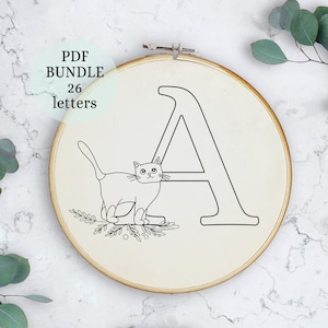 Puede incluir: Un dibujo en blanco y negro de un gato sentado junto a la letra A. La imagen está en un aro de bordado circular.