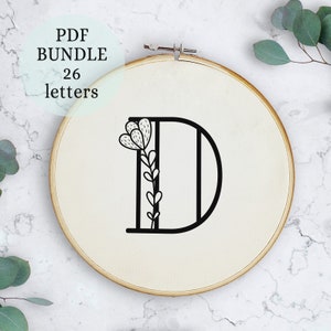 May include: A black and white embroidery pattern of the letter D with a floral design. The pattern is displayed in a wooden embroidery hoop.