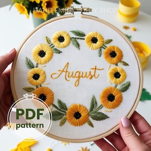 May include: A white embroidery hoop with a yellow and green floral design. The design features a wreath of sunflowers with green leaves and the word "August" in the center. The hoop is held by a person's hands.