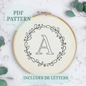 Puede incluir: Un patrón de bordado en blanco y negro de una corona con una letra A en el centro. El patrón incluye 26 letras y está disponible para descargar en PDF. "Incluye 26 letras" está escrito debajo de la corona.