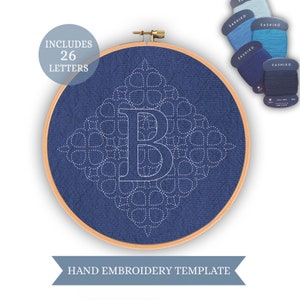 May include: A blue embroidery hoop with a white outline of the letter B in the center. The letter is surrounded by a geometric pattern of circles and squares. The hoop is labeled "HAND EMBROIDERY TEMPLATE" and "INCLUDES 26 LETTERS".